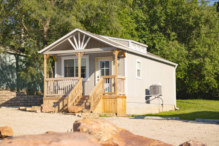 Swiss Cabin tan siding white trim set at edge of trees and grass